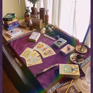 May include: A table set for a spiritual reading, featuring tarot cards, crystals, and a candle. The scene includes a purple velvet cloth, various decks of cards, and a small potted bonsai tree. A pencil and a book are also visible.