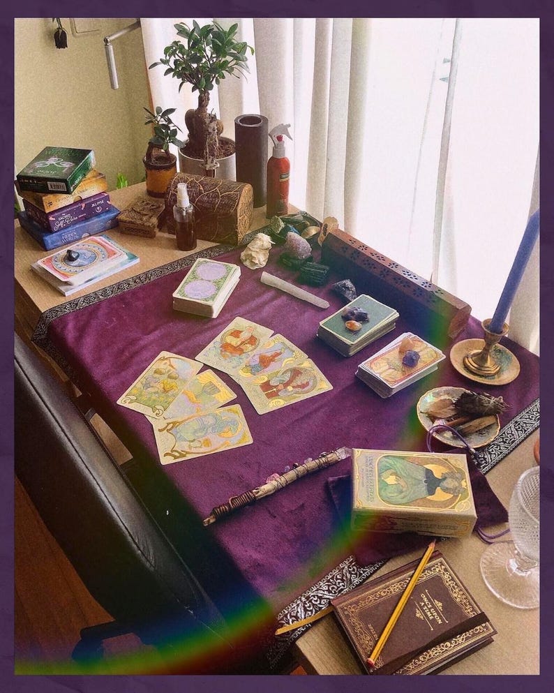 May include: A table set for a spiritual reading, featuring tarot cards, crystals, and a candle. The scene includes a purple velvet cloth, various decks of cards, and a small potted bonsai tree. A pencil and a book are also visible.