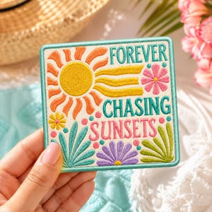 May include: Embroidered square patch with the words "FOREVER CHASING SUNSETS" in teal, pink, and yellow. The design includes a yellow sun, orange rays, and colorful flowers and leaves. The patch has a teal border.