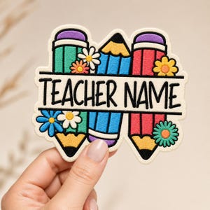 Può includere: Una toppa ricamata raffigurante tre matite colorate in verde acqua, blu e rosso con gomme viola. Piccoli fiori bianchi, arancioni e verde acqua sono sparsi tra le matite. Il testo TEACHER NAME è stampato in nero su un banner bianco.