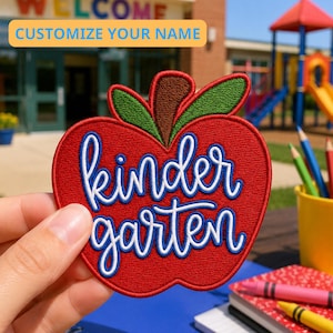 Puede incluir: Un parche bordado con forma de manzana, cuerpo rojo, tallo marrón y hojas verdes. La palabra kindergarten está cosida en letra cursiva blanca con un borde azul en el centro. Una pancarta amarilla en la parte superior dice CUSTOMIZE YOUR NAME.