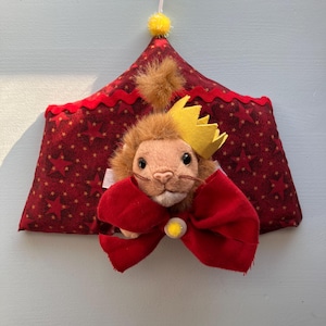 May include: A plush lion wearing a yellow crown and a red bow tie, set against a red fabric circus tent with gold star patterns. The tent has a red scalloped trim and a yellow pom-pom with a white flag.