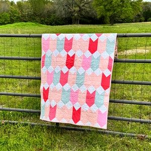 May include: A pink, red, and white quilt with a chevron pattern is hanging on a metal fence. The quilt is made of fabric squares sewn together to create a geometric design.