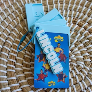 May include: A light blue luggage tag with the name "LINCOLN" in white letters. The tag is attached to a blue card featuring "The Wiggles" characters. The background is a woven basket.