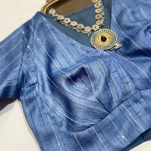 May include: A light blue blouse with vertical stripes and a V-neck. The blouse has short sleeves and is paired with a silver and gold necklace with a large pendant. The pendant has a dark red stone in the center.