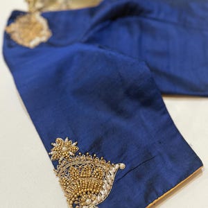 May include: A close-up of a navy blue blouse with intricate gold and pearl embroidery on the sleeve. The embroidery features floral and paisley designs. The blouse has a gold trim along the edges.