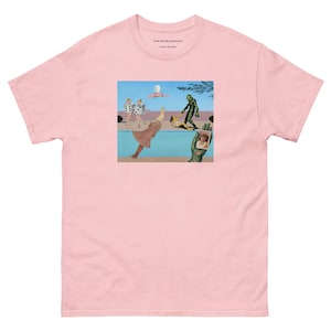 May include: Light pink t-shirt with a surrealist graphic print. The print features a collage of figures, including a green creature, a woman in a dress, and a hand holding a photo. The shirt has the text "The Big Long Uncomfortable Dance" and "Wilson Wheeler".