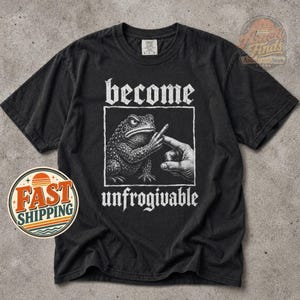 Può includere: Una t-shirt nera a maniche corte con una grafica bianca raffigurante un rospo e una mano umana che punta un dito. Il testo sopra la grafica recita "become" e il testo sottostante "unfrogivable" in un carattere bianco dall'aspetto vissuto.