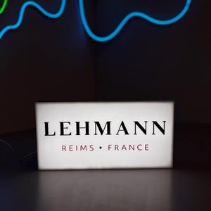 Op de afbeelding: Een rechthoekig, verlicht bord met de naam "LEHMANN" in vette zwarte letters. Daaronder, in kleinere rode letters, staat "REIMS • FRANCE". Het bord staat op een donkere ondergrond, met neonblauwe en groene lichten op de achtergrond.