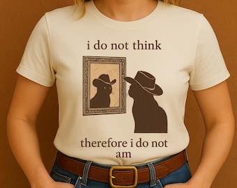 May include: Cream-colored t-shirt featuring a cat wearing a cowboy hat, looking at its reflection in a framed picture. The text "i do not think" and "therefore i do not am" are printed above and below the image.