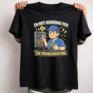 May include: Black t-shirt with a cartoon graphic of an electrician troubleshooting a panel. The text on the shirt reads "I'M NOT IGNORING YOU I'M TROUBLESHOOTING" in white and yellow. The electrician is wearing a blue uniform and cap.