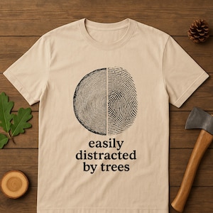Pode incluir: Uma camiseta bege com um gráfico preto. O gráfico é dividido verticalmente, com um design de anéis de árvore à esquerda e um design de impressão digital à direita. O texto "easily distracted by trees" está abaixo.
