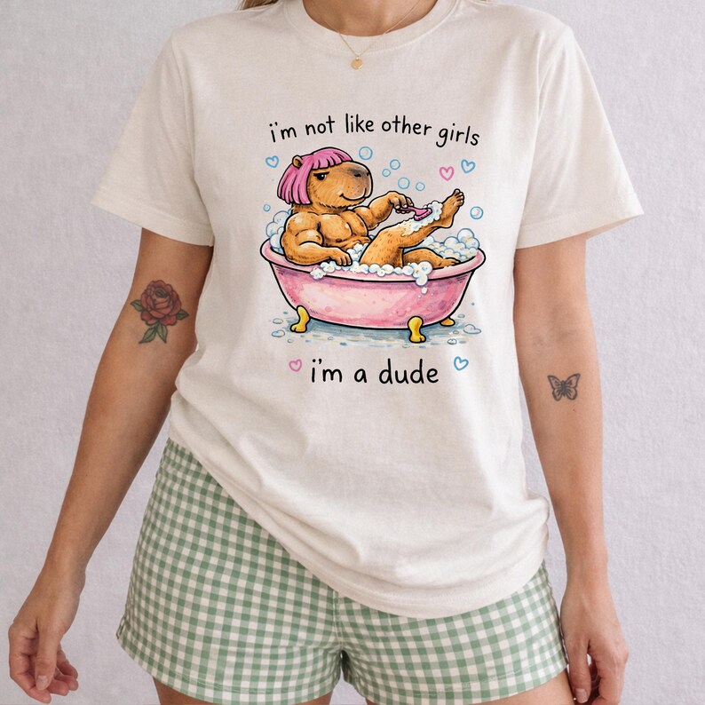 May include: Cream-colored t-shirt featuring a cartoon capybara in a pink bathtub, shaving its leg. The text "I'm not like other girls, I'm a dude" is printed above and below the illustration. The shirt is paired with green and white checkered shorts.