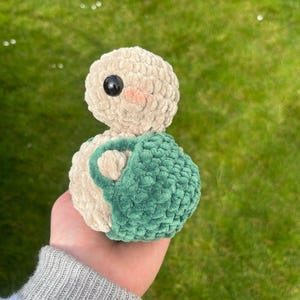 Pode incluir: Um brinquedo de pelúcia em crochê em forma de pássaro com uma bolsa verde em crochê. O pássaro tem um corpo bege com olhos pretos e bochechas cor-de-rosa.