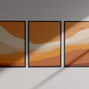 May include: Three framed abstract art prints with a sunset theme. The prints feature a layered landscape design in shades of orange, yellow, and brown. The text on the prints reads: "All the way lives on in me", "What remains toward remains alive", and "Carried along with the movement it becomes light".