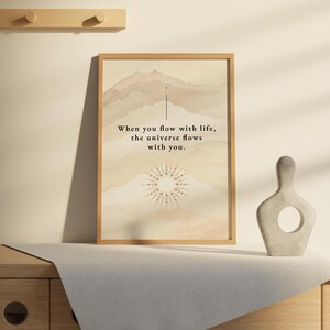 Op de afbeelding: Een ingelijste print met een beige achtergrond en een citaat dat luidt: "Wanneer je met het leven stroomt, stroomt het universum met je mee." De print toont een gestileerde zon met een bergketen op de achtergrond.