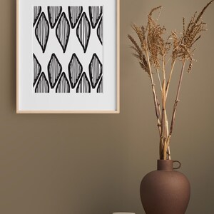 Op de afbeelding: Een ingelijst zwart-witdruk van een geometrisch patroon hangt aan een lichtbruine muur. Een vaas met gedroogde grassen staat op een houten tafel met een kleine ronde container.