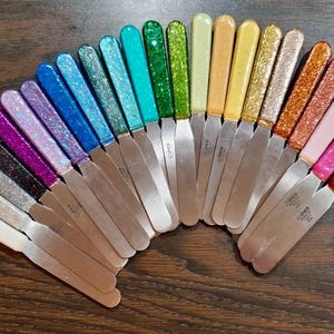 May include: A collection of colorful palette knives with glitter handles. The handles are in a rainbow of colors, including white, black, purple, blue, green, yellow, orange, pink, and red. The stainless steel blades are arranged in a fan shape.