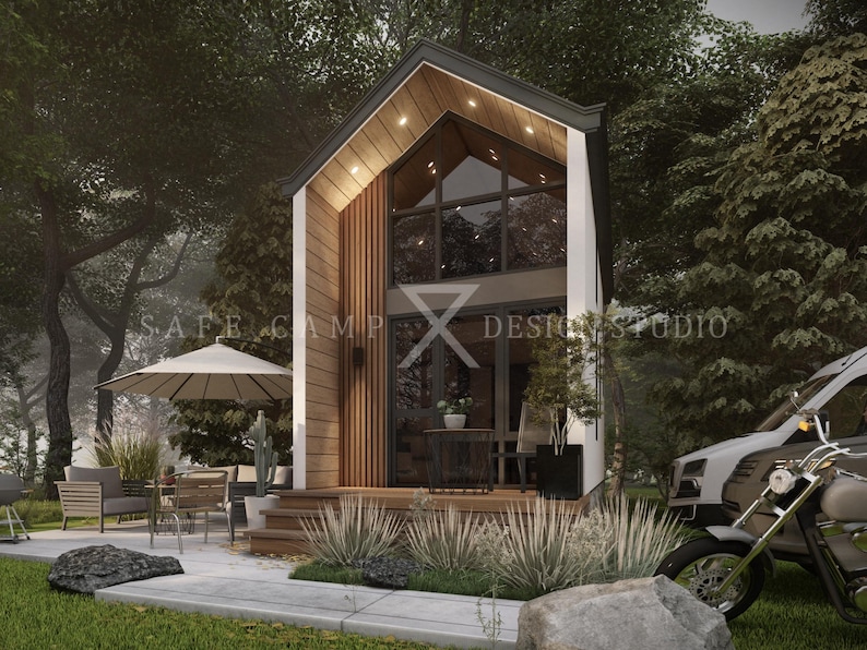 May include: A modern tiny house with a high-pitched roof, large windows, and wooden accents. The house has a small porch with outdoor furniture and a motorcycle parked nearby. The text "SAFE CAMP Design Studio" is visible.