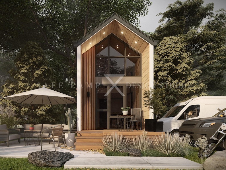 May include: A modern tiny house with a large glass window and wooden accents. The house has a small porch with a table and chairs, and is surrounded by trees and landscaping. The text "SAFE CAMP DESIGN STUDIO" is visible.