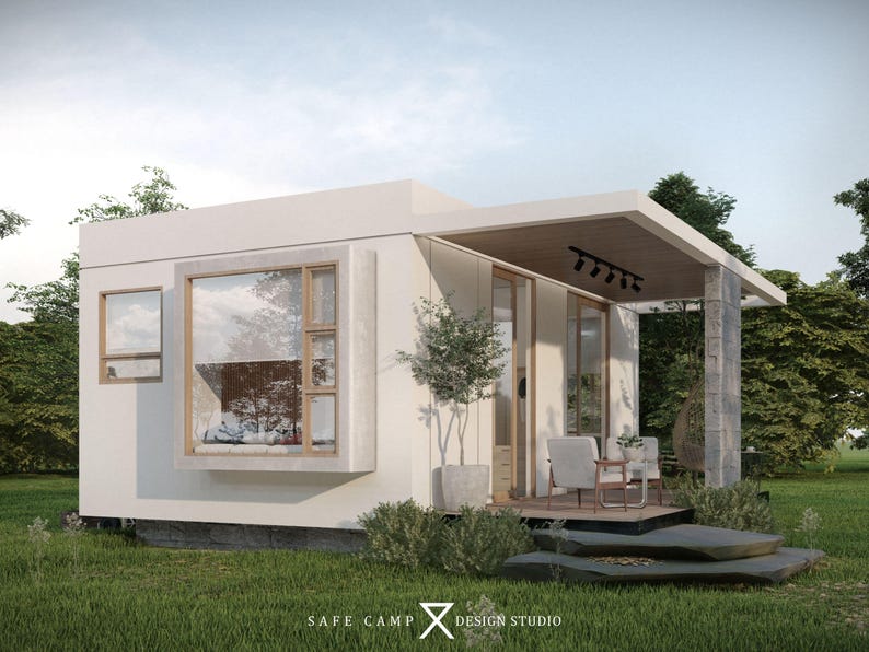 May include: A modern, white cabin with large windows and a covered porch. The cabin features a minimalist design with a flat roof and is surrounded by green grass and trees. The text "SAFE CAMP DESIGN STUDIO" is visible.