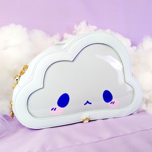 May include: White cloud-shaped handbag with a clear window. The bag features a cartoon face with blue eyes, pink cheeks, and a small black mouth. A gold chain strap and a small gold ring are attached to the bag.