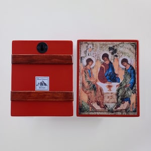 May include: A red wooden icon with a religious image. The back of the icon is red with two brown wooden strips and a black hook. The front features a detailed depiction of three figures in robes, with halos, seated around a table.