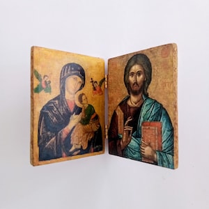 Puede incluir: Un icono religioso de madera con bisagras que presenta dos paneles. Un panel representa a María y el niño Jesús, el otro a Jesús sosteniendo un libro. La obra de arte tiene un fondo dorado y marrón con detalles en azul, verde y rojo.
