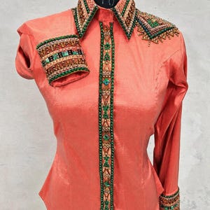 Peut inclure: Une chemise à manches longues de couleur corail avec des ornements complexes sur le col, les poignets et la patte de boutonnage. La bordure est décorée de rangées de petites perles vertes, d'accents bronze et de cristaux verts carrés.