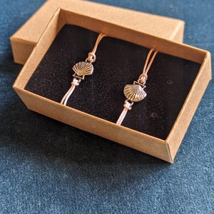 May include: Two delicate pink string bracelets with silver seashell charms in a brown box.