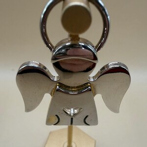 May include: A silver-colored angel-shaped keychain. The keychain features a detailed design of an angel with outstretched wings. The angel has a heart-shaped detail on its chest and a ring for attaching keys. The keychain is made of a shiny, reflective metal.