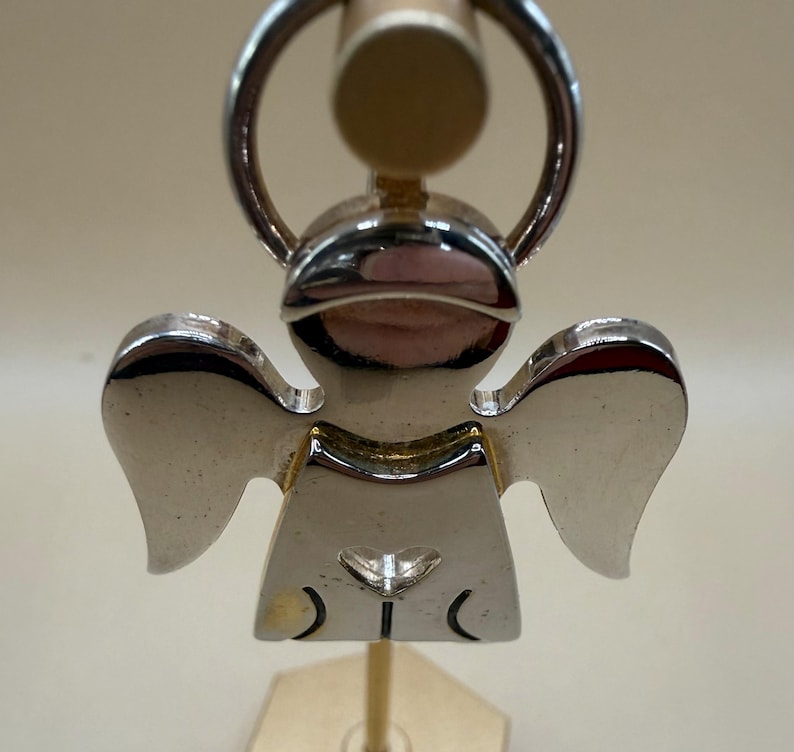 May include: A silver-colored angel-shaped keychain. The keychain features a detailed design of an angel with outstretched wings. The angel has a heart-shaped detail on its chest and a ring for attaching keys. The keychain is made of a shiny, reflective metal.