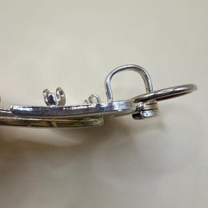 May include: Close-up of a silver-toned metal earring with a curved design and a small loop. The earring has a shiny finish and appears to be made of a durable metal. The design includes a circular element and a small, decorative detail.