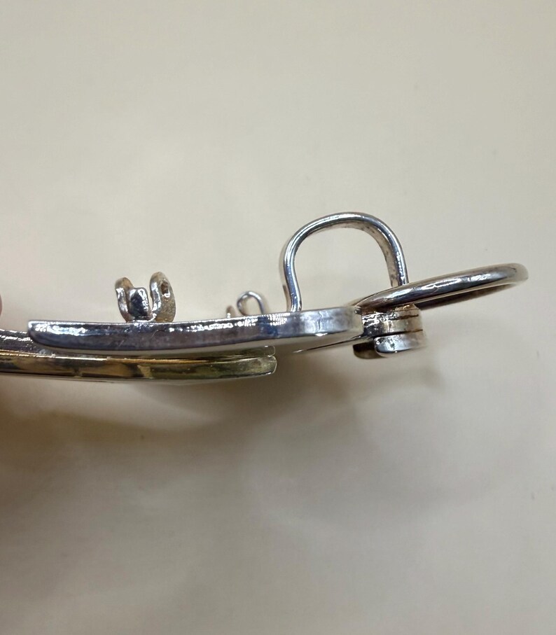 May include: Close-up of a silver-toned metal earring with a curved design and a small loop. The earring has a shiny finish and appears to be made of a durable metal. The design includes a circular element and a small, decorative detail.