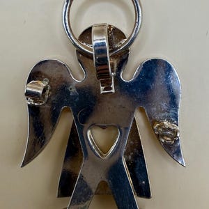 May include: A silver-colored angel pendant with a heart-shaped cutout and a ring at the top. The angel has outstretched wings and is designed with a simple, stylized form. The pendant is likely made of metal.