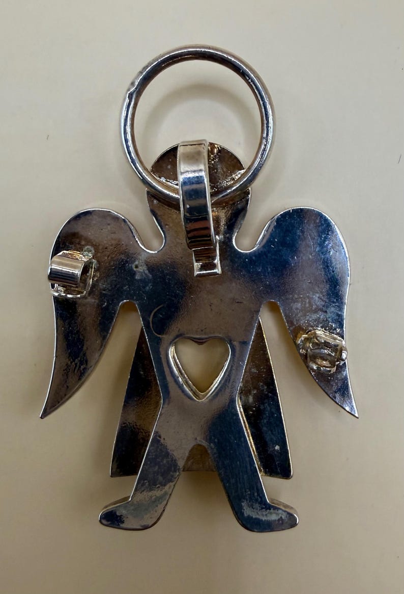 May include: A silver-colored angel pendant with a heart-shaped cutout and a ring at the top. The angel has outstretched wings and is designed with a simple, stylized form. The pendant is likely made of metal.