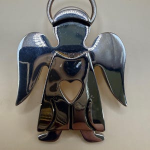 May include: A shiny silver angel-shaped keychain with a halo and heart-shaped cutout. The angel has outstretched wings and a reflective surface, creating a bright and eye-catching design. The keychain is made of metal.