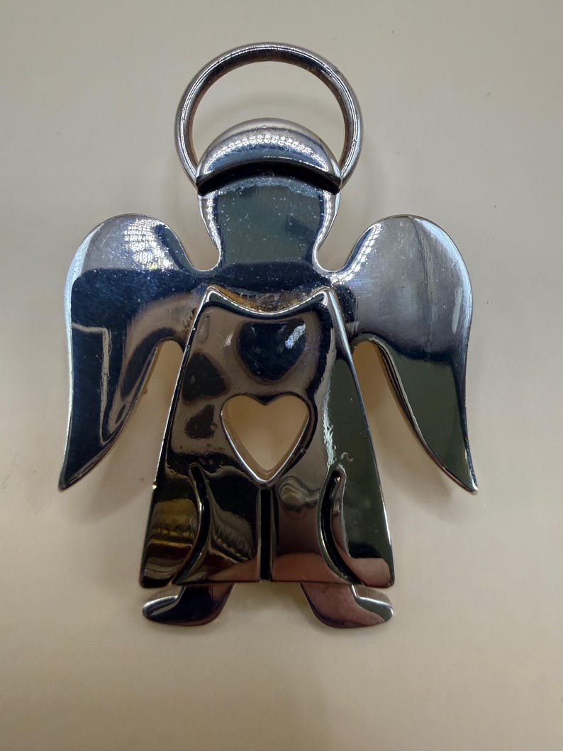 May include: A shiny silver angel-shaped keychain with a halo and heart-shaped cutout. The angel has outstretched wings and a reflective surface, creating a bright and eye-catching design. The keychain is made of metal.