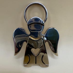 May include: A silver-colored angel-shaped keychain with a halo and heart-shaped cutout. The angel has outstretched wings and a simple, stylized design. The keychain is made of a shiny, reflective metal.