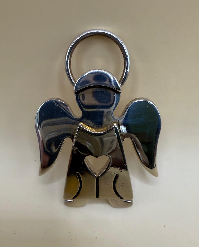 May include: A silver-colored angel-shaped keychain with a halo and heart-shaped cutout. The angel has outstretched wings and a simple, stylized design. The keychain is made of a shiny, reflective metal.