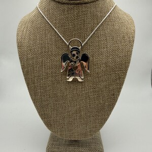 May include: A silver-toned angel pendant necklace. The pendant features an angel with outstretched wings and a heart-shaped detail on the chest. The necklace is displayed on a brown mannequin.