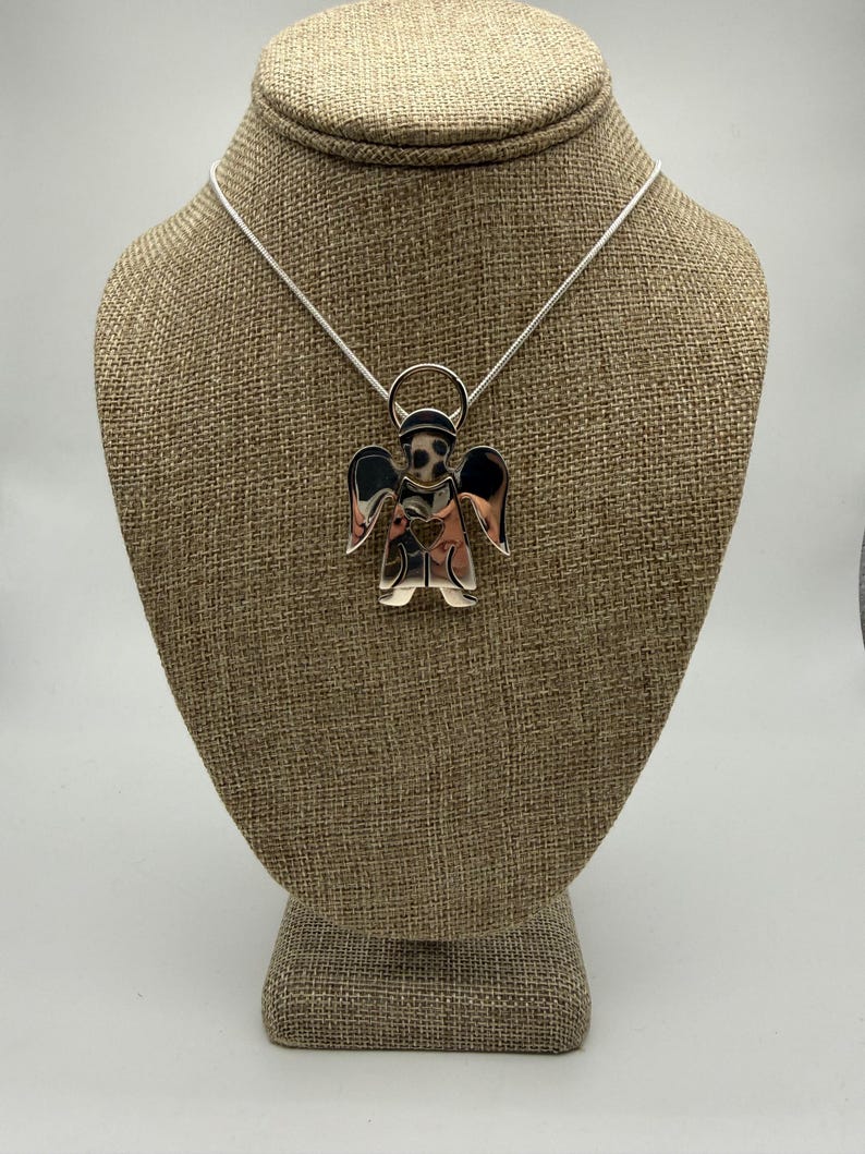 May include: A silver-toned angel pendant necklace. The pendant features an angel with outstretched wings and a heart-shaped detail on the chest. The necklace is displayed on a brown mannequin.