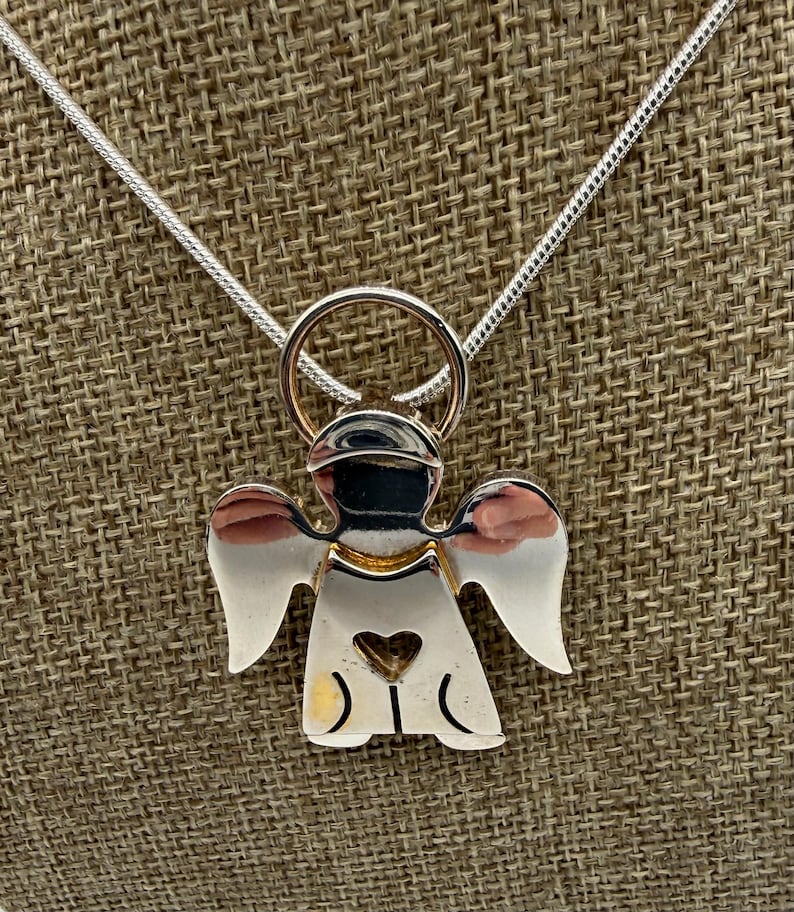 May include: A silver angel pendant necklace with a heart cutout. The angel has outstretched wings and a halo. The necklace chain is a thin, silver snake chain. The pendant is made of polished silver and gold-colored metal.