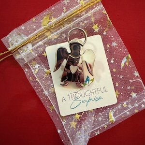 May include: A silver angel pin with a heart detail, presented on a card that reads "A Thoughtful Surprise." The pin is displayed in a sheer, star-patterned gift bag with a gold drawstring, set against a red background.