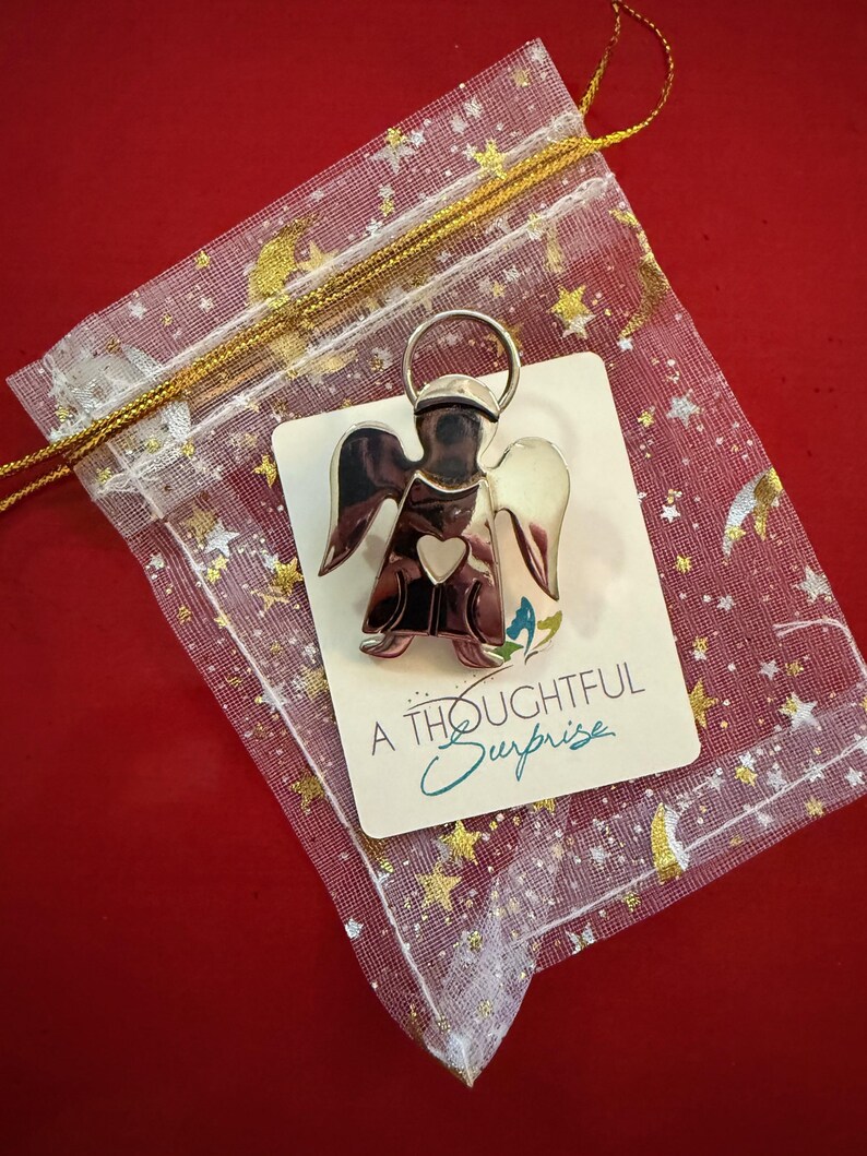 May include: A silver angel pin with a heart detail, presented on a card that reads "A Thoughtful Surprise." The pin is displayed in a sheer, star-patterned gift bag with a gold drawstring, set against a red background.