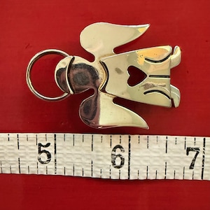 May include: A silver-toned metal angel-shaped keychain with a heart-shaped cutout. The keychain is approximately 2 inches long, with a ring for attaching keys. The background is a solid red color.