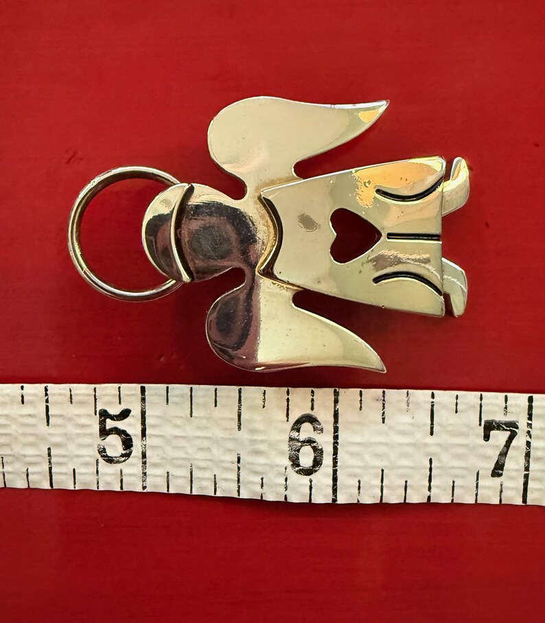 May include: A silver-toned metal angel-shaped keychain with a heart-shaped cutout. The keychain is approximately 2 inches long, with a ring for attaching keys. The background is a solid red color.