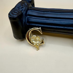 May include: A gold-colored brooch in the shape of the letter "C" with a small cherub figure seated inside. The brooch has a polished finish and is set against a black background.