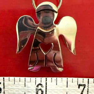 May include: A gold-toned angel pendant with a heart-shaped cutout. The angel has outstretched wings and a halo. The pendant is approximately 2 inches in height. The background is a solid red color.