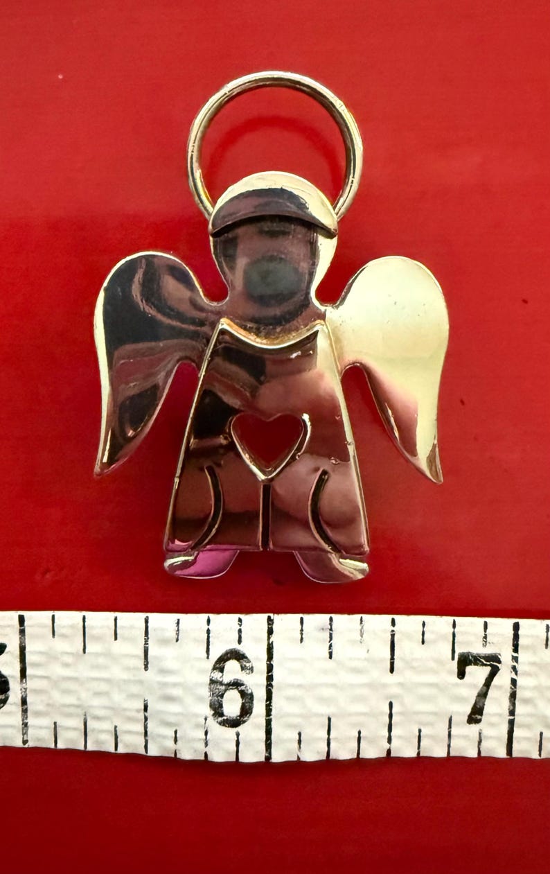 May include: A gold-toned angel pendant with a heart-shaped cutout. The angel has outstretched wings and a halo. The pendant is approximately 2 inches in height. The background is a solid red color.